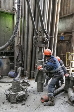 İki sondaj işçisi sondaj ipinin altını söküyor. Spiral kalibratörü söküyorum. Bir makine anahtarıyla çalışmak. Sondaj kulesinin döner platformunda bulunuyorlar..