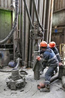 İki sondaj işçisi sondaj ipinin altını söküyor. Spiral kalibratörü söküyorum. Bir makine anahtarıyla çalışmak. Sondaj kulesinin döner platformunda bulunuyorlar..