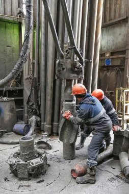 İki sondaj işçisi sondaj ipinin altını söküyor. Spiral kalibratörü söküyorum. Bir makine anahtarıyla çalışmak. Sondaj kulesinin döner platformunda bulunuyorlar..