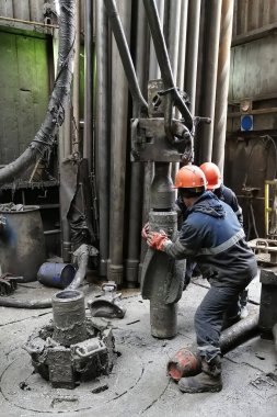 İki sondaj işçisi sondaj ipinin altını söküyor. Spiral kalibratörü söküyorum. Bir makine anahtarıyla çalışmak. Sondaj kulesinin döner platformunda bulunuyorlar..