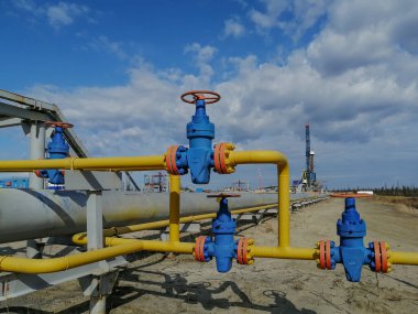 Boru hattı tesisatları ve sahada gaz kuyuları üretme manifoltları. Yüksek basınçlı valfler için el tekerlekleri. Arka planda bir sondaj kulesi var. Bulanık uzak nesneler.