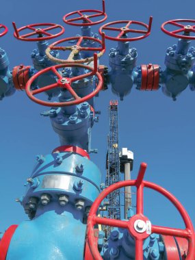Yüksek basınçlı kapı valfları olan bir gaz kuyusu çeşmesi. Sondaj kulesi arka planda. Kompozisyonun bazı ayrıntıları bulanık. Yaz günü, mavi gökyüzü.