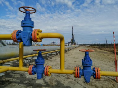 Boru hattı tesisatları ve sahada gaz kuyuları üretme manifoltları. Yüksek basınçlı valfler için el tekerlekleri. Arka planda bir sondaj kulesi var. Bulanık uzak nesneler.