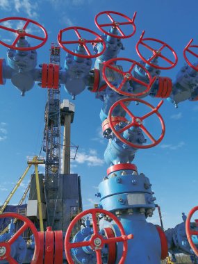 Yüksek basınçlı kapı valfları olan bir gaz kuyusu çeşmesi. Sondaj kulesi arka planda. Kompozisyonun bazı ayrıntıları bulanık. Yaz günü, mavi gökyüzü.