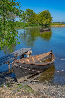 Güneşli bir yaz gününde Onega Gölü 'ndeki Kizhi adasında manzara. Öndeki gölün yanında büyük ahşap bir tekne. Arka planda gölde yüzen bir tekne, ağaçlar ve göl genişliyor.