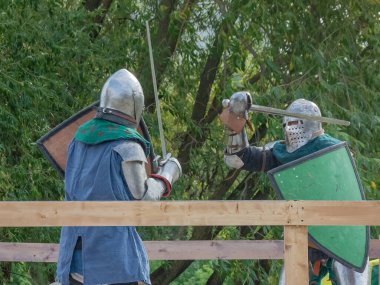 Ağır ortaçağ zırhlı iki şövalye kılıçlarla savaşıyor. Demir miğferler ve kalkanlar tarafından korunuyorlar. Ortaçağ Avrupa şövalye turnuvalarının tarihi yeniden yapılandırması