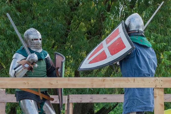 Two knights on foot in heavy medieval armor fight with swords. They are protected by iron helmets and shields. Historical reconstruction of medieval European knightly tournaments