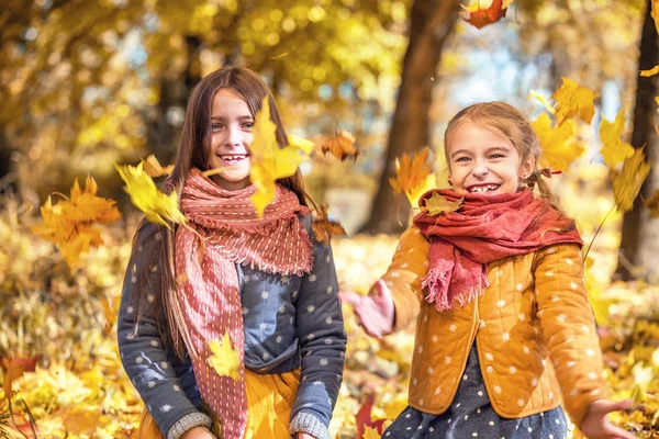 Zwei süße lächelnde 8-jährige Mädchen, die an einem sonnigen Herbsttag in einem Park mit Blättern spielen. — Stockfoto