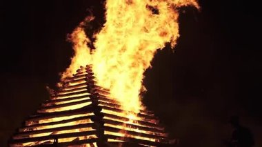 Büyük, ahşap bir piramidin geceleri alev alev yanan dramatik yavaş çekimi. Uzun alevler karanlık gökyüzüne karşı kükrer, kıvılcımlar ve parlayan közler uçuşur. Açık hava yaz şenlik ateşi, siluetli bir seyirci ile..