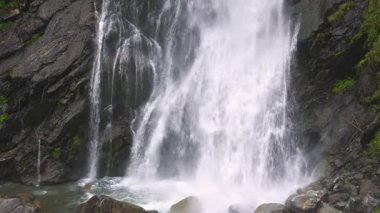 Bir dağ şelalesinin yavaş çekimde karanlık kayalara çarpıp berrak bir havuza çarpması. Sprey, sis ve köpük ferahlatıcı, ıslak ve huzurlu bir doğa sahnesi yaratır.. 