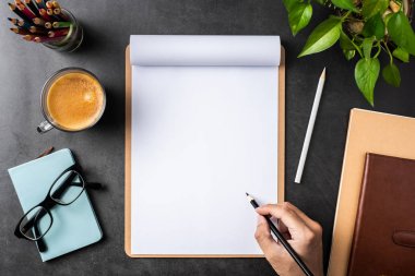 Spiral defter, saksı bitki, gözlük, kahve, kalem ve siyah arka plan üzerinde çanta ile erkek el üst görünümü.