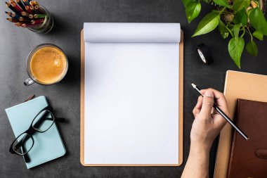 Spiral defter, saksı bitki, gözlük, kahve, kalem ve siyah arka plan üzerinde çanta ile erkek el üst görünümü.