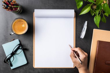 Spiral defter, saksı bitki, gözlük, kahve, kalem ve siyah arka plan üzerinde çanta ile erkek el üst görünümü.