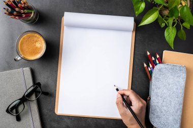 Spiral defter, saksı bitki, gözlük, kahve, kalem ve siyah arka plan üzerinde çanta ile erkek el üst görünümü.