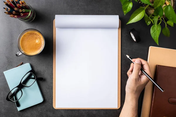 Spiral defter, saksı bitki, gözlük, kahve, kalem ve siyah arka plan üzerinde çanta ile erkek el üst görünümü.