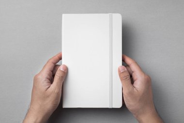 Gri arka planda beyaz defter tutan erkek ellerin üst görünümü.