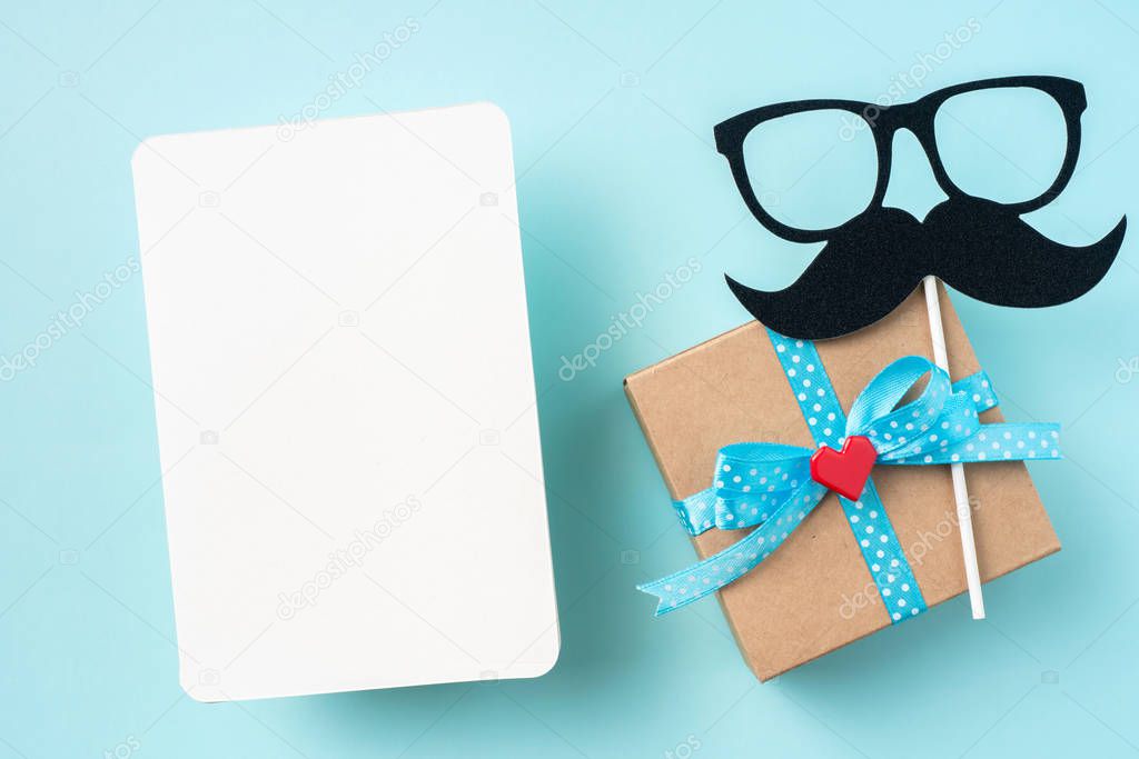 Top view of fathers day layout with gift box, square greeting card, silhouette of eyeglasses and mustache.