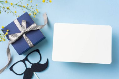 Beyaz tebrik kartı, gümüş kurdele kraft hediye kutusu, sarı çiçekler, gözlük, mavi arka plan bıyık ile Düzen.