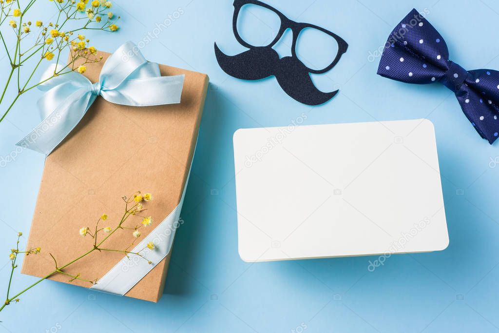 Fathers day layout with ribbon kraft gift box, yellow flowers, necktie, eyeglasses and mustache on blue background.