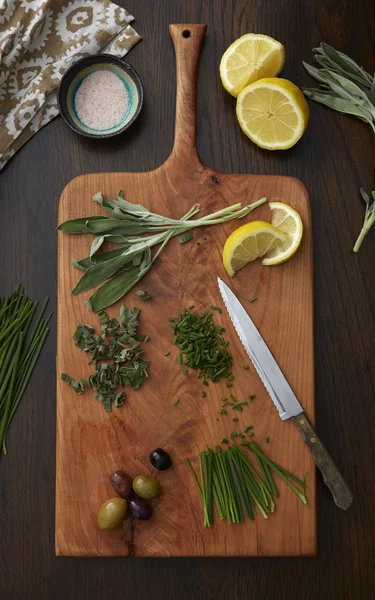 Herbs, olives, and lemons on a wood cutting board with a knife, napkin and bowl of salt on a wood table