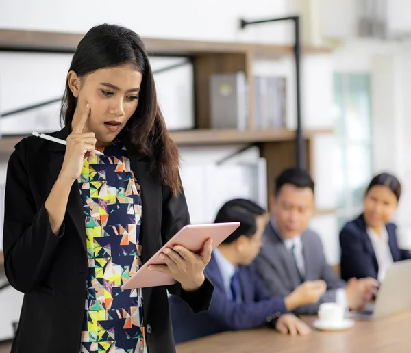 Portrait of confidence asian Businesswoman working and thinking with smart tablet and stand in front of table in meeting room of cafe with business team in background using for coporate background work