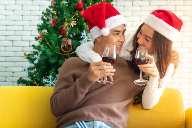 Genç Asyalı ergen çift Noel tatilini birlikte şarap içerek kutluyor oturma odasında Noel ağacı süslemesiyle..
