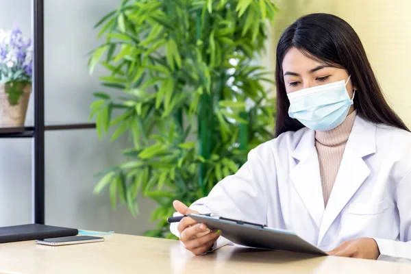 アジアの女性医師の肖像画は 保護顔マスクを着用し 病院の診療所の彼女のオフィスルームに座って 忍耐を確認する前に忍耐ファイルを読んでください ヘルスケアの概念 — ストック写真