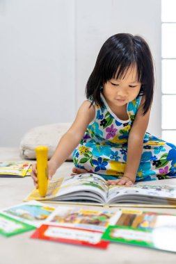 Asian girl child reading interactive book in living room at home as home schooling while city lockdown because of covid-19 pandemic across the world. Home Schooling prepare for Preschool concept. clipart