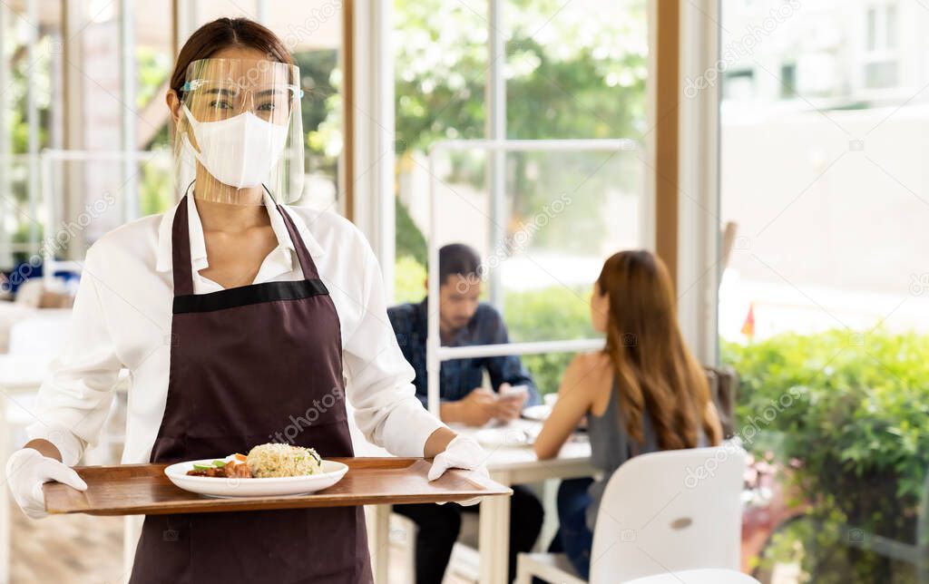 Portrait attractive Asian waitress wear face mask and face shield holding food tray to serving meal to customer with customers in background. New normal restaurant concept.