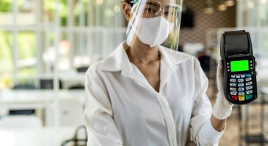 Portre çekici Asyalı garson yüz maskesi takıyor ve yüz maskesi takıyor. Kapalı restoran geçmişi olan bir kredi kartı okuyucusu için kredi kartı okuyucu tutuyor. Yeni normal restoran ulaşılamaz ödeme konsepti.