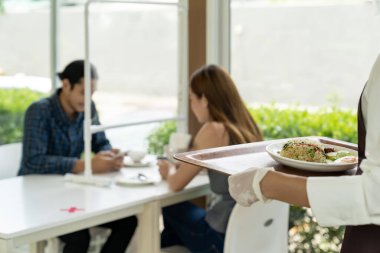 Arkadan bakan Asyalı garson, arka planda müşterisi olan müşterilere yemek servisi için yemek tepsisi tutuyor. Yeni normal restoran yaşam tarzı konsepti