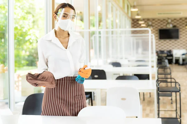 Portrait attractive asian waitress wear face mask and face shield hold alcohol and wet wipe for cleaning table before welcome customer. New normal hygiene restaurant concept.