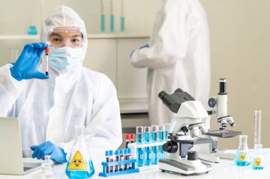 Bilim adamları kan örneği test tüpünü tutuyor ve araştırması için inceliyor ve arka planda meslektaşı ile birlikte Coronavirus covid-19 salgını aşısı geliştiriyor. Tıp bilimi ve teknoloji konsepti.