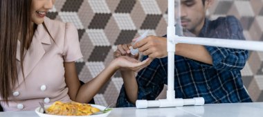 Panoramik Asyalı adam, yeni normal bir sosyal servis restoranında yemek yemeden önce ortağına alkol jeli dezenfektanı sürmüş. Restoran, yeni normal yaşam tarzı. Web pankartı mahsulü.