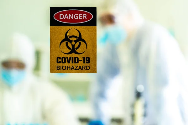 Caution danger Signage of COVID-19 coronavirus in front of Laboratory room with background of Scientists working and researching vaccine for ncov-19 virus for world pandemic situation.