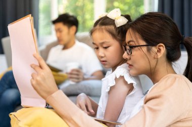 Asyalı kız, annesiyle oturma odasında babasıyla masal kitabı okurken Coronavirus covid-19 salgınından dolayı şehir kilitlenirken evde çalışıyor. Aile hayatı ve ev konsepti..