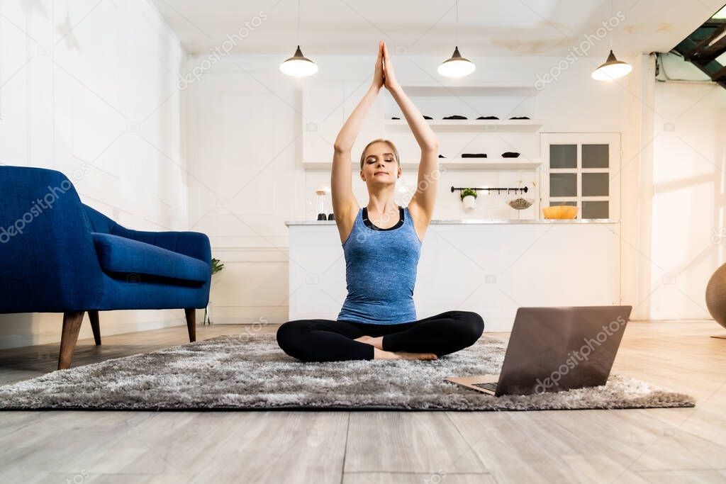 White caucasian fit sporty woman doing yoga Sukhasana sit pose in living room at home watching online tutorials from trainer on laptop. Working out, fitness sport and healthy lifestyle concept.