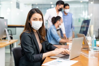 Asyalı ofis çalışanı iş kadını portresi yeni normal ofiste koruyucu yüz maskesi takıyor ve sosyal uzaklık uygulaması Coronavirus COVID-19 'u engellerken ırklar arası takım arka planda yer alıyor..