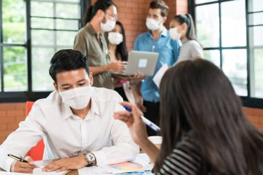 İki iş adamı, arka planda çalışan ırklar arası iş adamları grubuyla toplantı odasında beyin fırtınası yapıyor. Yeni normal ofiste yüz maskesi takıyorlar. Koronavirüs COVID 19 yayılmasını önlüyorlar.