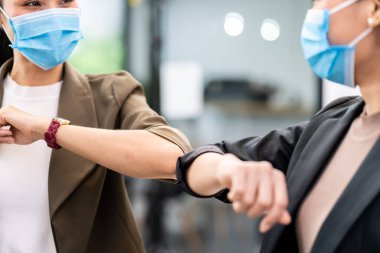 İki iş kadını yeni normal ofis hayatı tarzında alternatif karşılama yapıyorlar. Korunaklı yüz maskesi takıyorlar ve Coronavirus COVID-19 enfeksiyonunu azaltmak için el sıkışmak yerine dirsek teması kullanıyorlar..