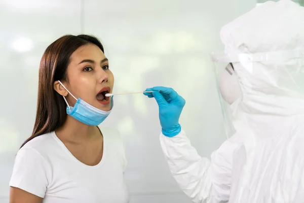 Medical staff with PPE suit test coronavirus covid-19 to asian woman by throat swab at home. New normal health care service at home and medical delivery and COVID-19 testing concept.