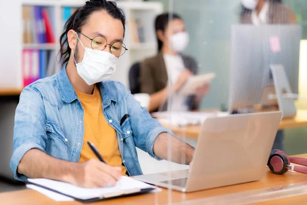 Asian office employee businessman wear protective face mask work in new normal office with interracial colleague in background as social distance practice prevent coronavirus COVID-19.