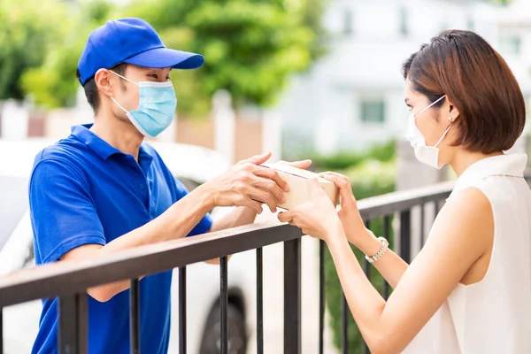 アジア人は青いシャツを着た男に荷物を渡し 家の若い女性の衣装を与えます 彼らは コロナウイルスCovid 19拡散を減らすためにフェイスマスクを着用します パッケージショッピング配信コンセプト — ストック写真