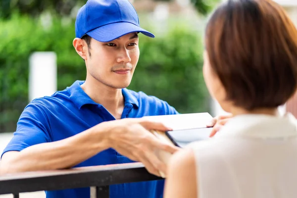 Asian deliver man handling package box to asian woman in front of house and female customer sign electronic signature to portable mobile device. Package shopping delivery and digital sign concept.