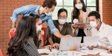 Asyalı beyaz iş adamlarından oluşan panoramik bir grup toplantı ve toplantı odasında tekrar tekrar beyin fırtınası yapıyorlar. Koronavirüs enfeksiyonunu önlemek için koruyucu yüz maskesi takıyorlar..