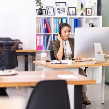 Yetişkin, arkadaş canlısı, kulaklıklı Asyalı bir kadın sosyal uzaklık masası olan bir çağrı merkezinde çalışıyor. Çağrı merkezi ve tele market satış konsepti.