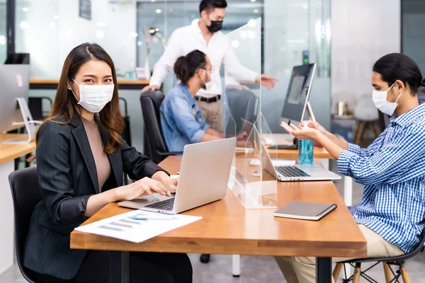 Portrait of asian office employee businesswoman wear protective face mask work in new normal office with interracial team in background as social distance practice prevent coronavirus COVID-19.