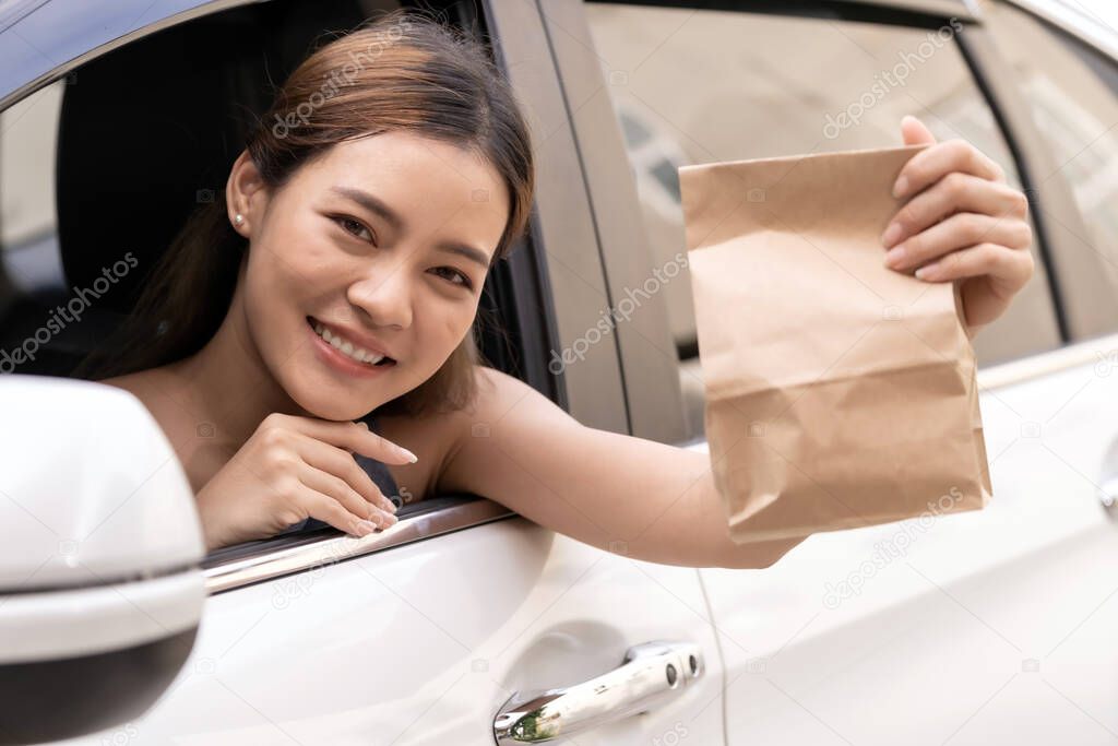 Asian Young adult in car holding disposable bag for take out food from drive thru service restaurant. Drive thru is new normal and popular service after coronavirus covid-19 pandemic.