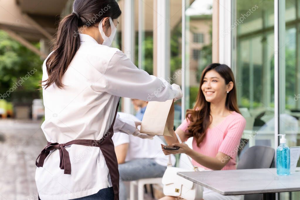 Back view of asian waitress with face mask give order of take out food bag to attractive woman female customer. Take away or take-out food service concept in new normal after coronavirus pandemic.
