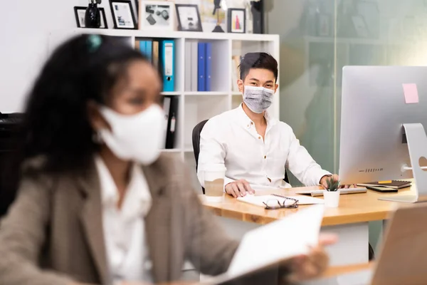 Portrait asian Office employee with protective face mask working in new normal office with social distance practice to american african colleague in foreground prevent coronavirus COVID-19 spreading.